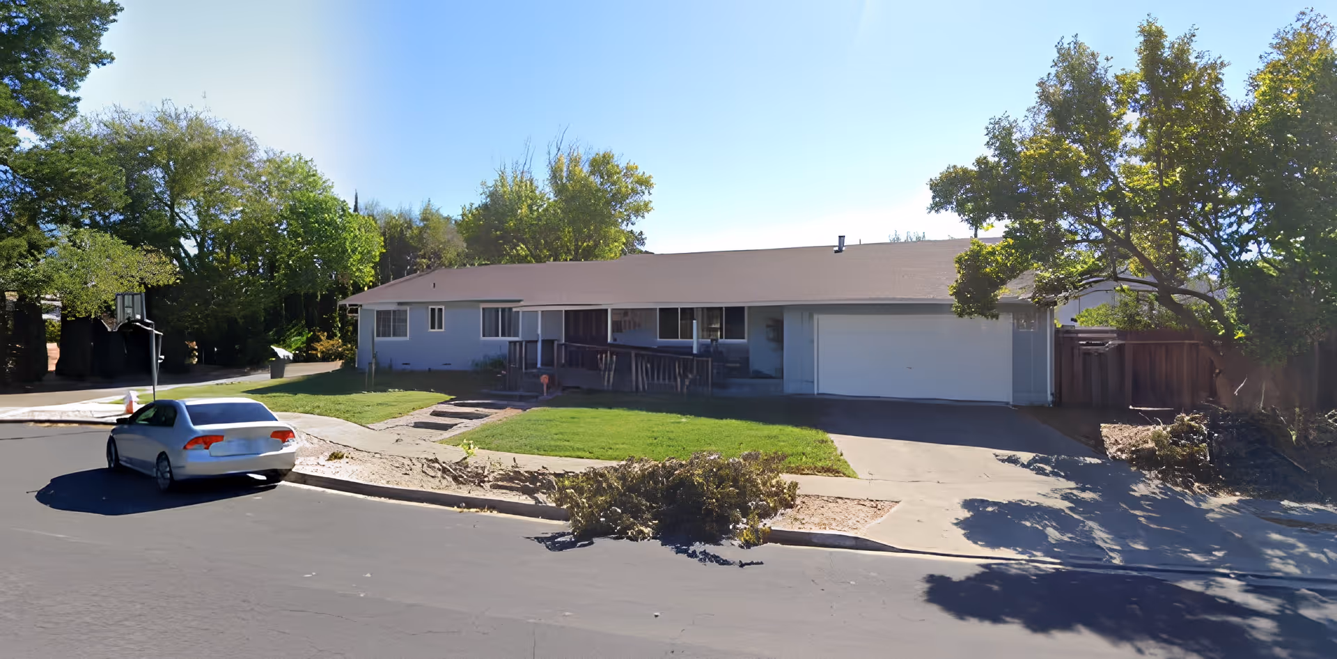 Single-story residential building with a light-colored exterior, a sloped roof, and a front porch with a wheelchair ramp. There is a green lawn in front, a driveway leading to a garage, and a silver car parked on the street. Trees surround the property under a clear blue sky.