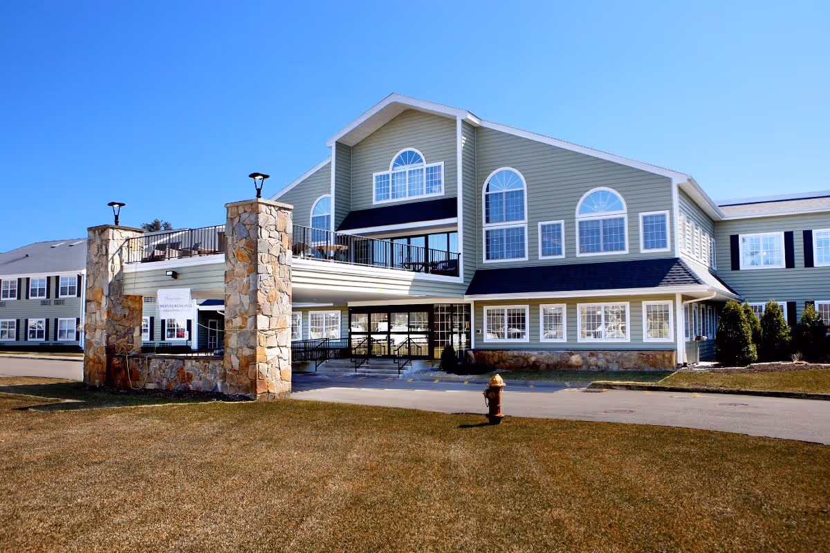 Front exterior of a large two-story senior living facility with stone pillars, covered entrance, and arched windows.