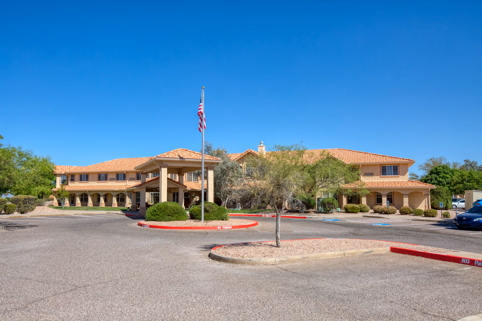 Exterior view of Vista Pointe at Sierra Vista, a two-story building with a tiled roof and covered entrance. The building is surrounded by a parking lot with some landscaping including bushes and small trees. An American flag is flying on a flagpole near the entrance under a clear blue sky.