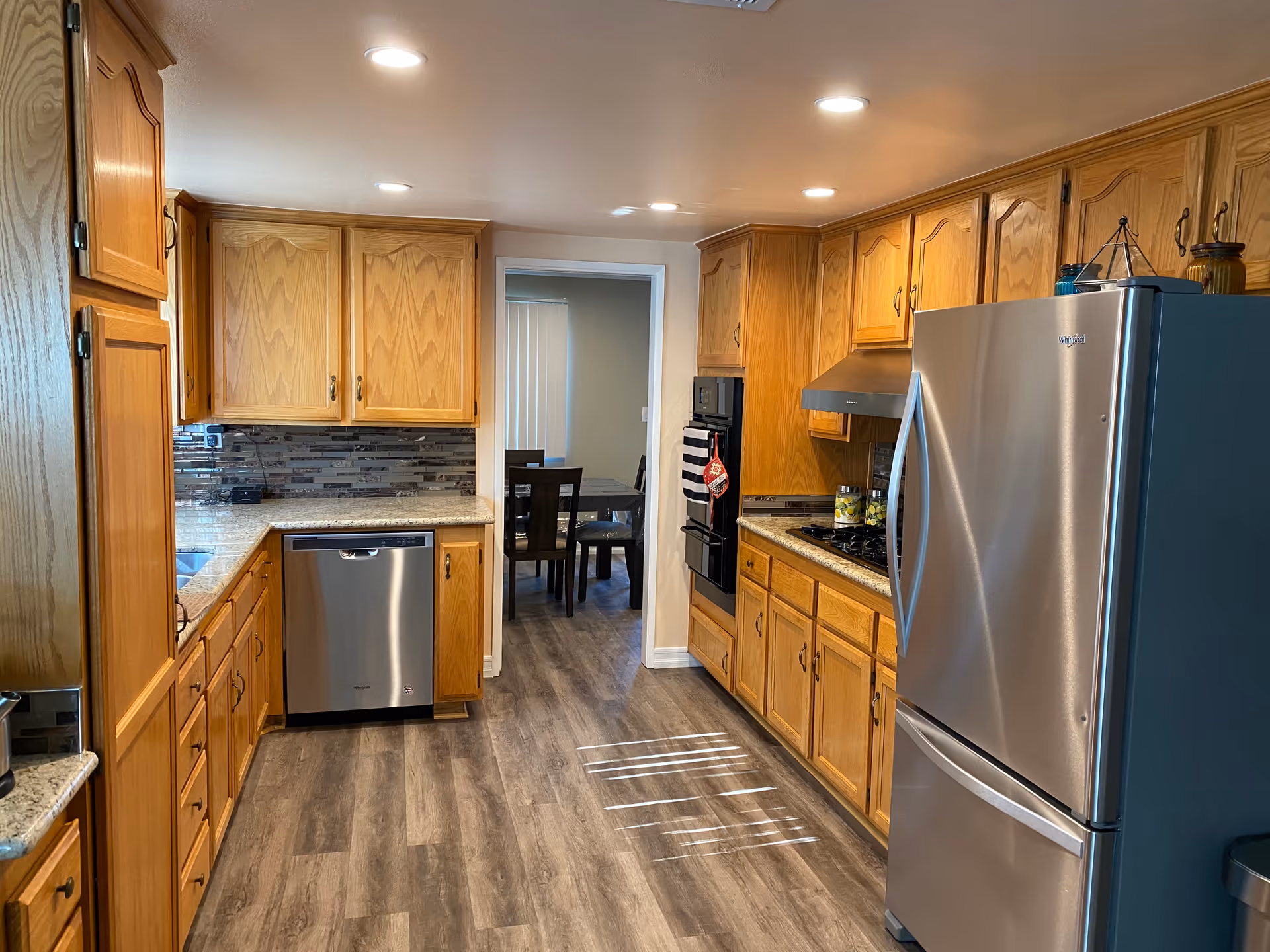 A modern kitchen with wooden cabinets, granite countertops, and stainless steel appliances including a refrigerator, dishwasher, oven, and stove. The kitchen has recessed lighting and wood-style flooring. Through a doorway, a dining area with a table and chairs is visible.