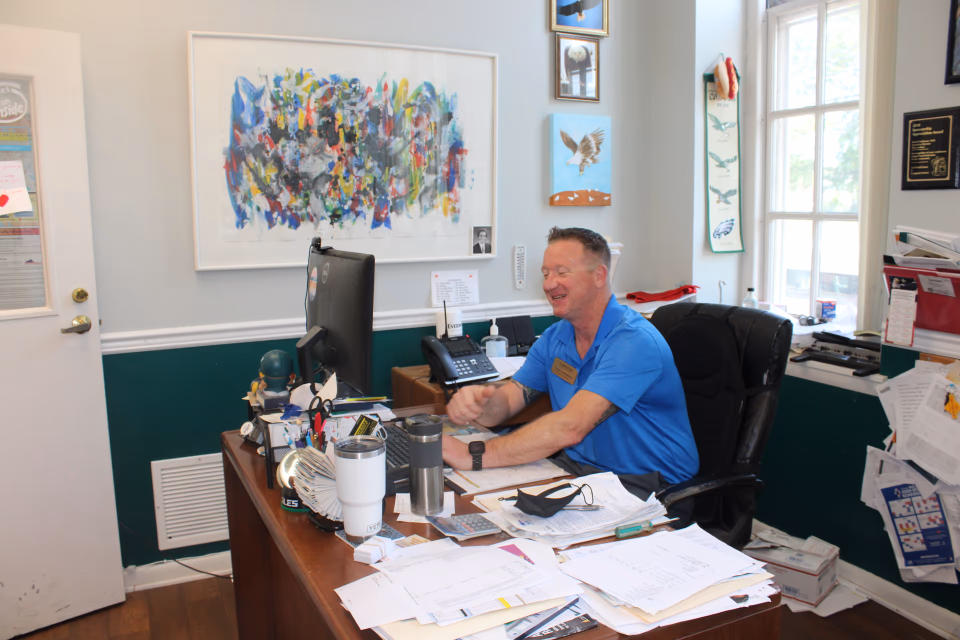 A man in a blue shirt sitting at a cluttered desk in an office, smiling and interacting with someone off-camera. The office has a large window, various papers and office supplies on the desk, and colorful artwork on the walls.