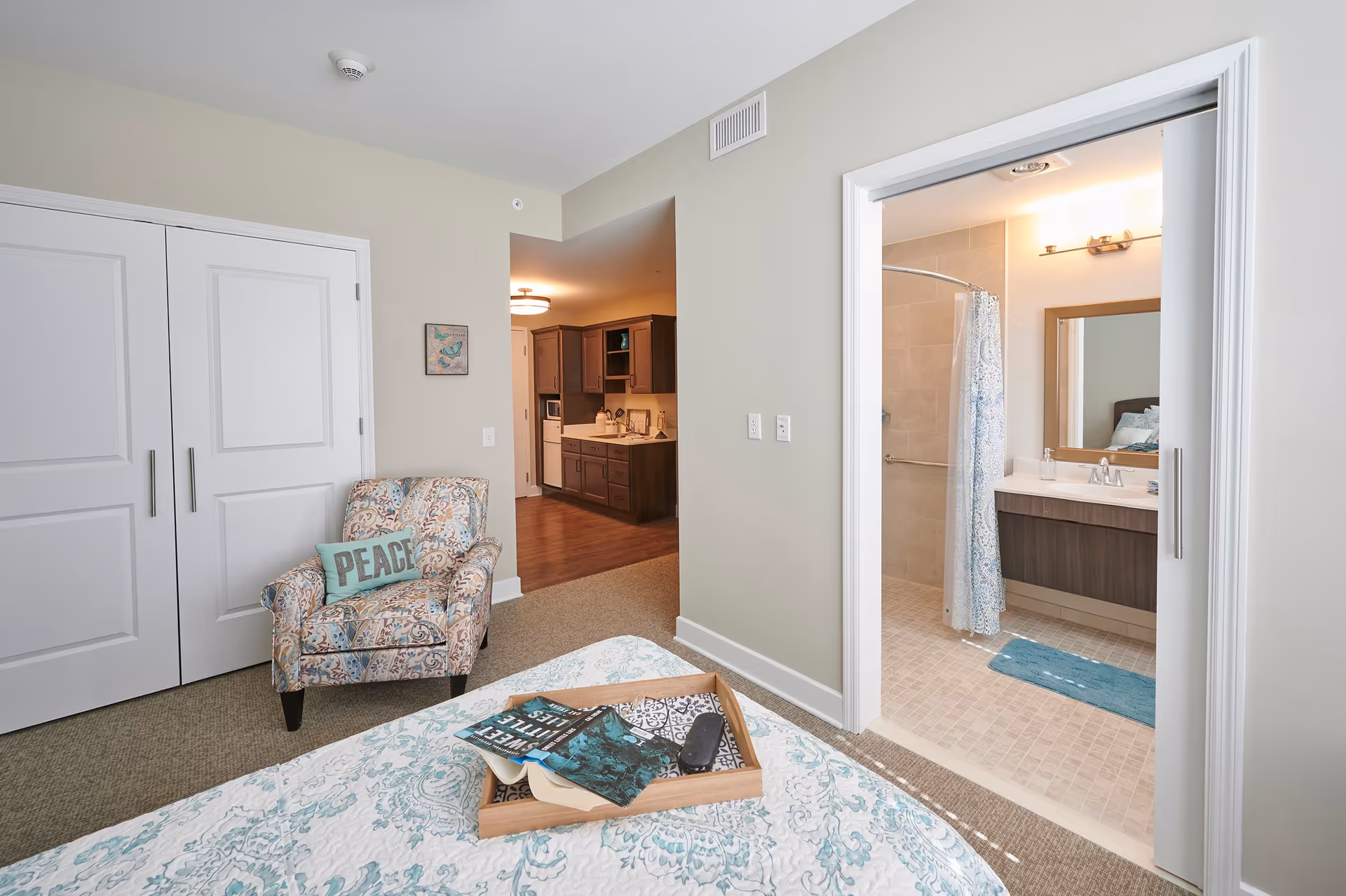 A cozy bedroom with a patterned armchair holding a pillow that says 'PEACE', a bed with a tray containing books and a remote, and an open door leading to a bathroom with a shower and a vanity with a mirror. The kitchen area is visible in the background.