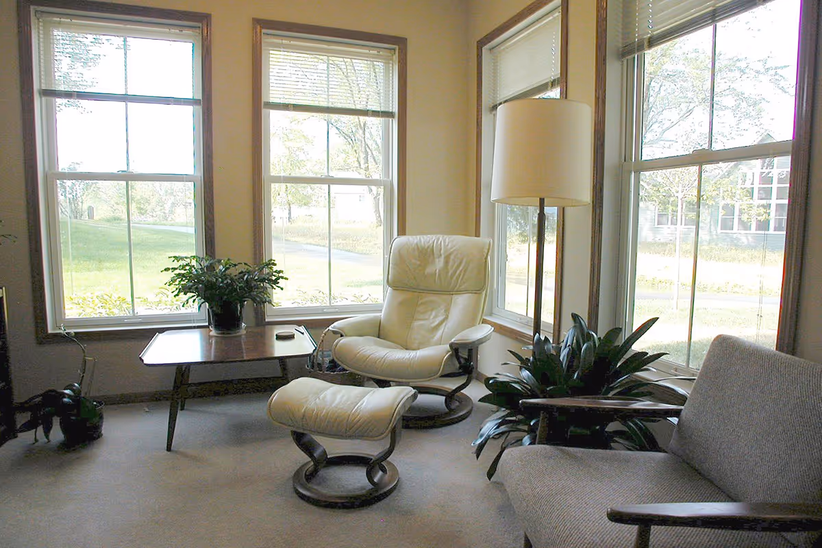 A cozy sitting area with large windows letting in natural light. The room features a white leather armchair with a matching ottoman, a wooden side table with a potted plant, a floor lamp, and a gray upholstered chair. Green plants are placed near the windows, and the view outside shows grass and trees.