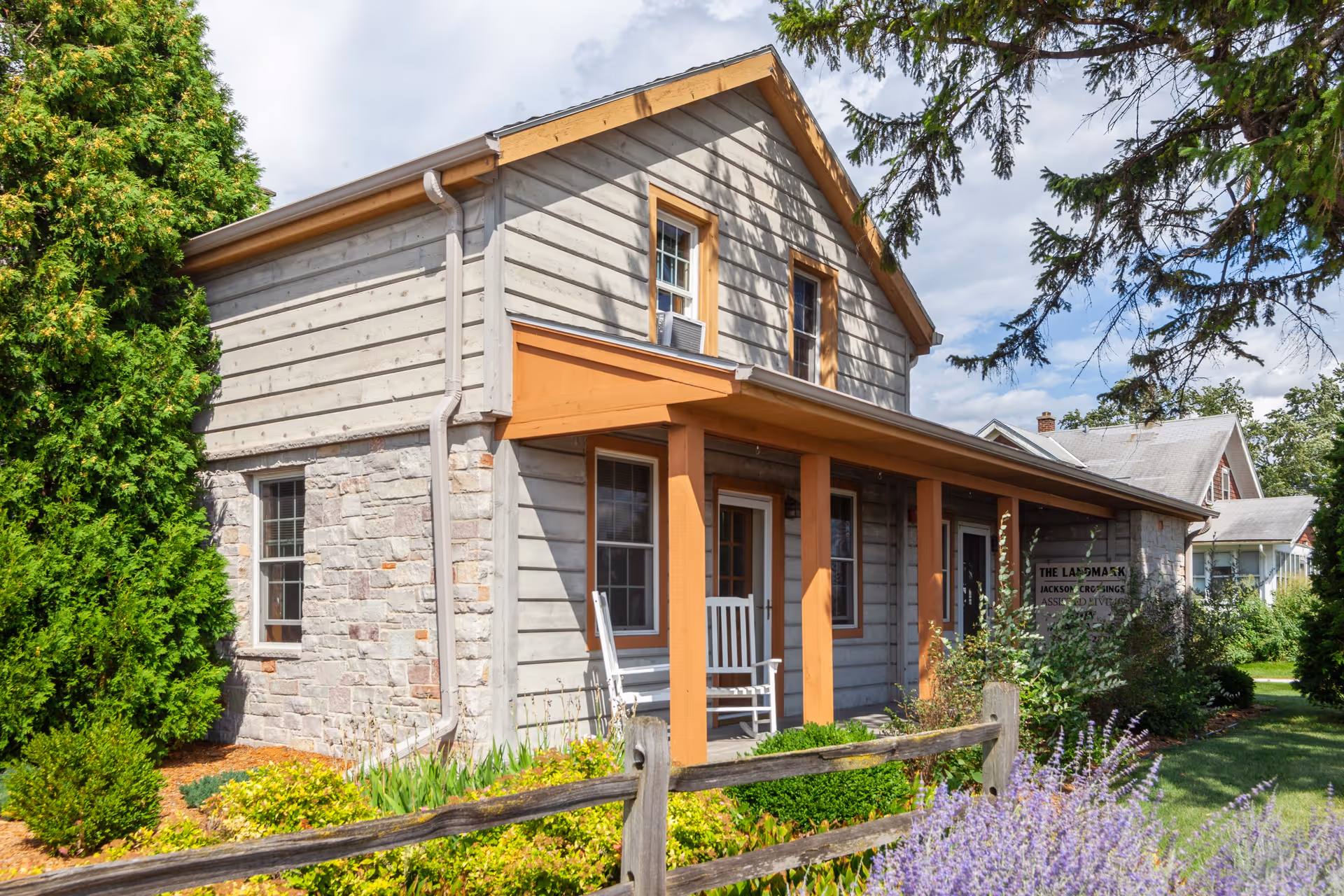 Exterior view of a two-story senior living facility building with a stone and wood facade, a covered porch with a white rocking chair, surrounded by greenery and flowers under a partly cloudy sky.