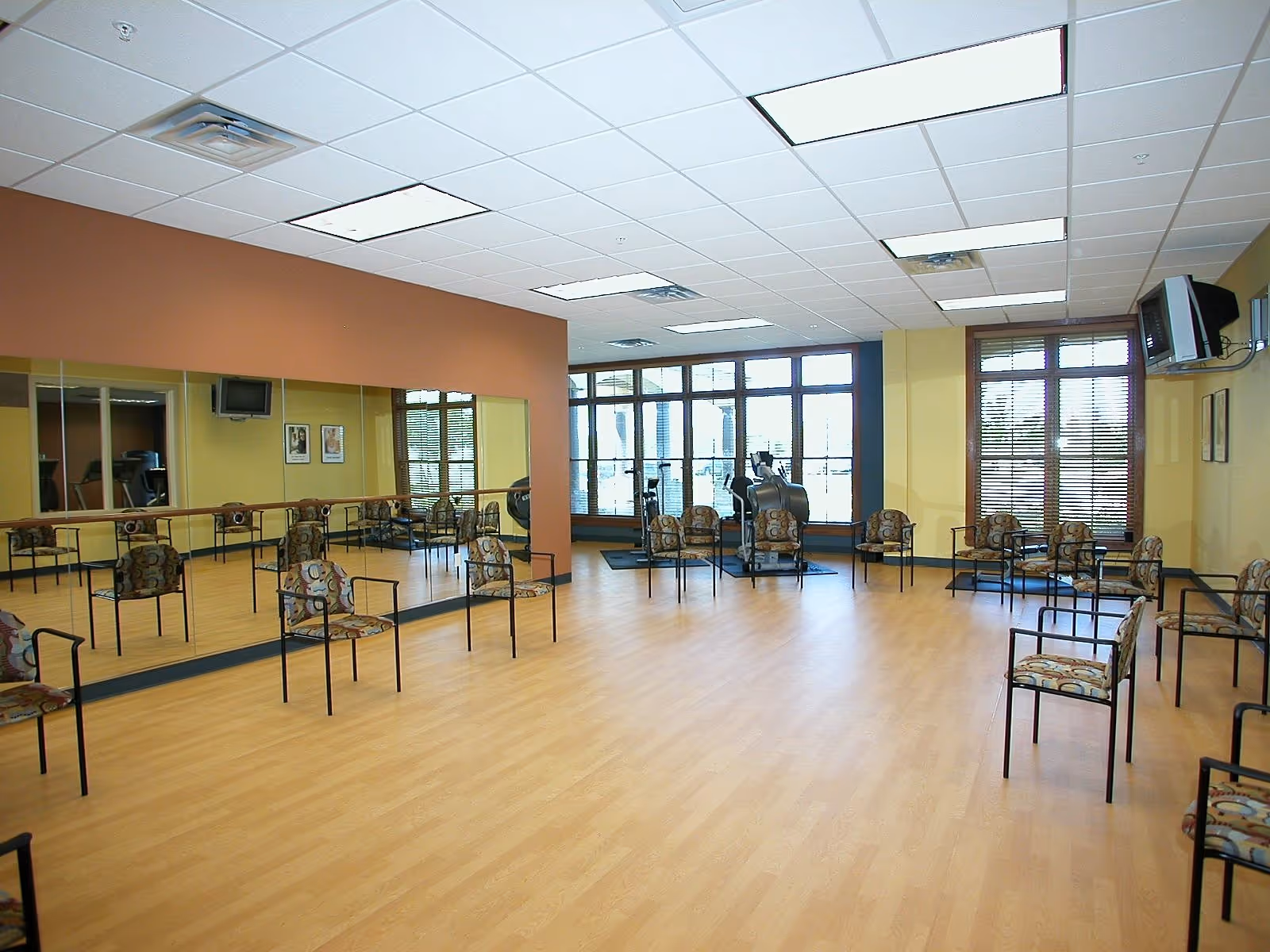 A spacious exercise or activity room with wooden flooring, multiple patterned chairs arranged around the perimeter, large mirrors on one wall, windows with blinds, and exercise equipment near the windows. The room has a drop ceiling with fluorescent lights and a wall-mounted TV.