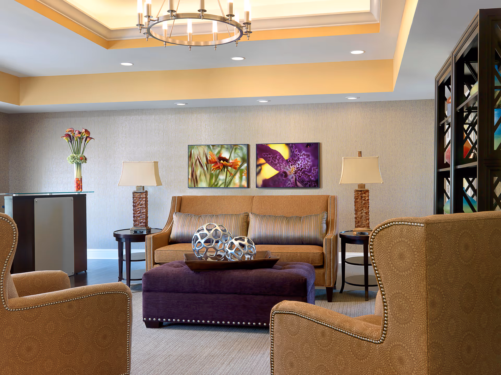 A cozy living room area with a brown sofa, two matching armchairs, and a purple ottoman in the center. The sofa has two striped cushions, and there are two side tables with lamps on either side. On the wall above the sofa are two framed floral artworks. A modern chandelier hangs from the ceiling, and a tall vase with flowers is placed on a small cabinet to the left.