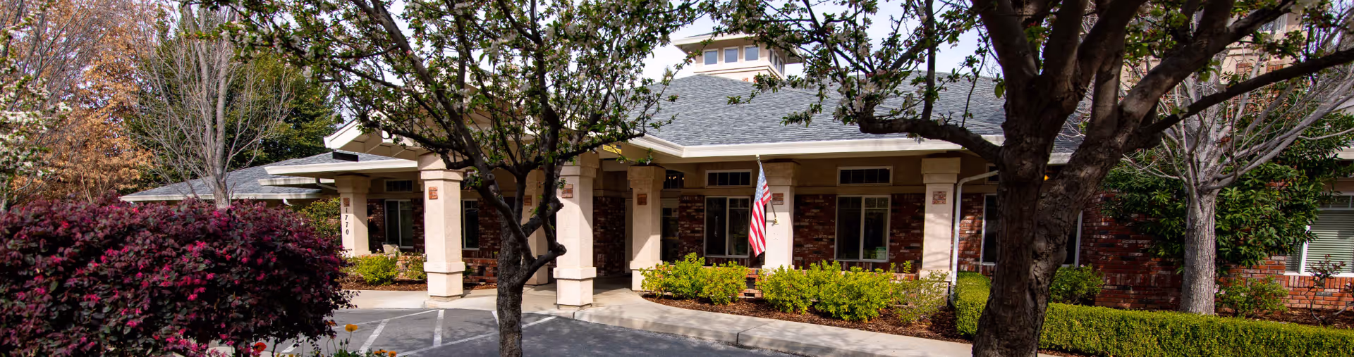 Exterior view of a senior living facility named Courtyard At Little Chico Creek, showing a single-story building with a covered entrance, brick and beige walls, surrounded by trees and shrubs, with an American flag displayed near the entrance.