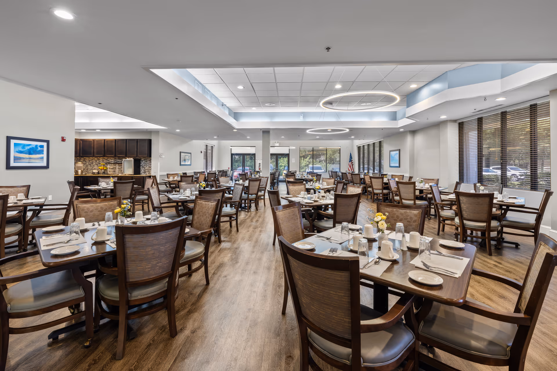 A spacious senior living dining room with multiple wooden tables and chairs arranged neatly. Each table is set with white plates, cups, glasses, and silverware. The room has large windows with blinds, modern circular ceiling lights, and a wooden floor. There is a small kitchen area visible in the background with dark cabinets and a tiled backsplash.