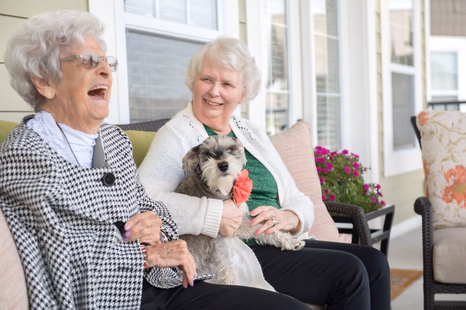Two elderly women sitting on a porch bench, one laughing and the other smiling while holding a small dog with a red flower collar. There are flowers and cushioned chairs in the background.