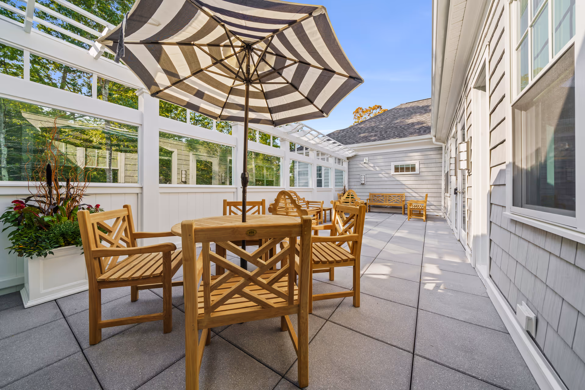 Outdoor patio area with wooden chairs and tables, a large striped umbrella, potted plants, and a white fence with glass panels. The patio is adjacent to a gray building with windows and doors, under a clear blue sky.