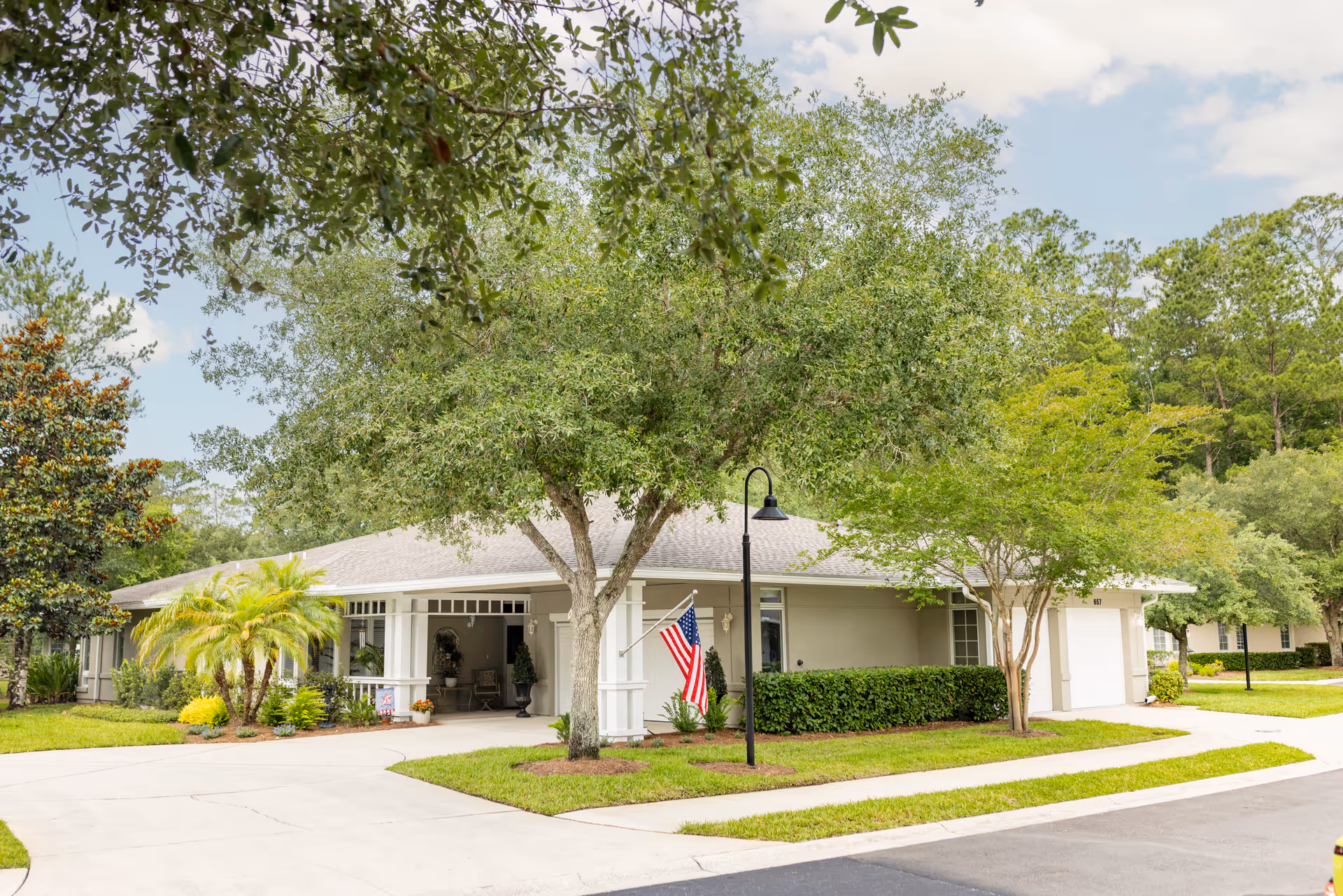 Exterior view of a single-story building at Westminster St. Augustine with a light gray roof, surrounded by green trees and shrubs. An American flag is displayed near the entrance, and a black street lamp stands on the sidewalk in front of the building.