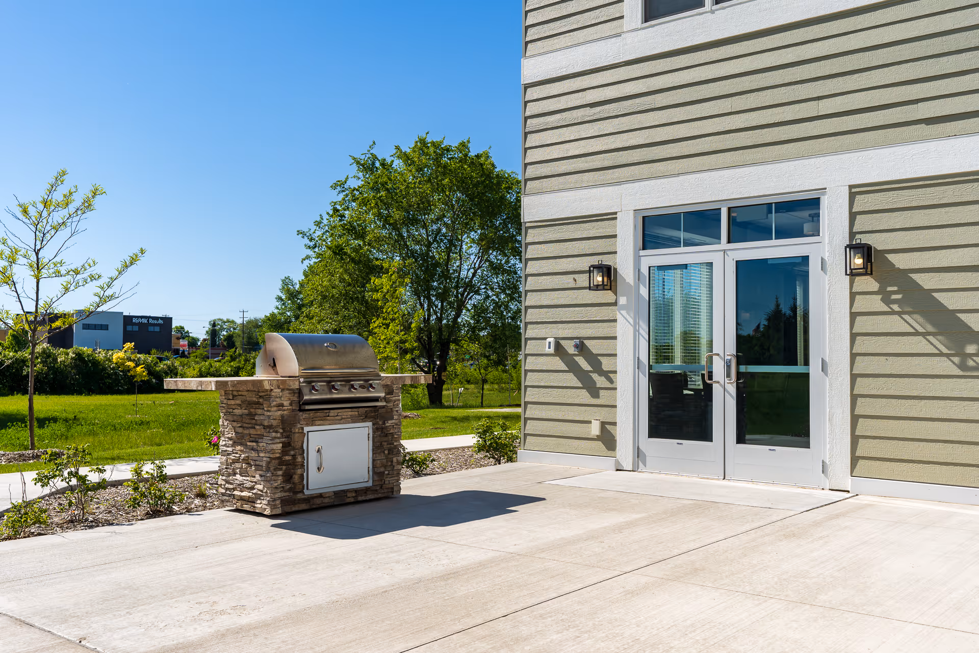 Outdoor patio with a built-in stone grill beside glass double doors on a light-green building under a clear blue sky.
