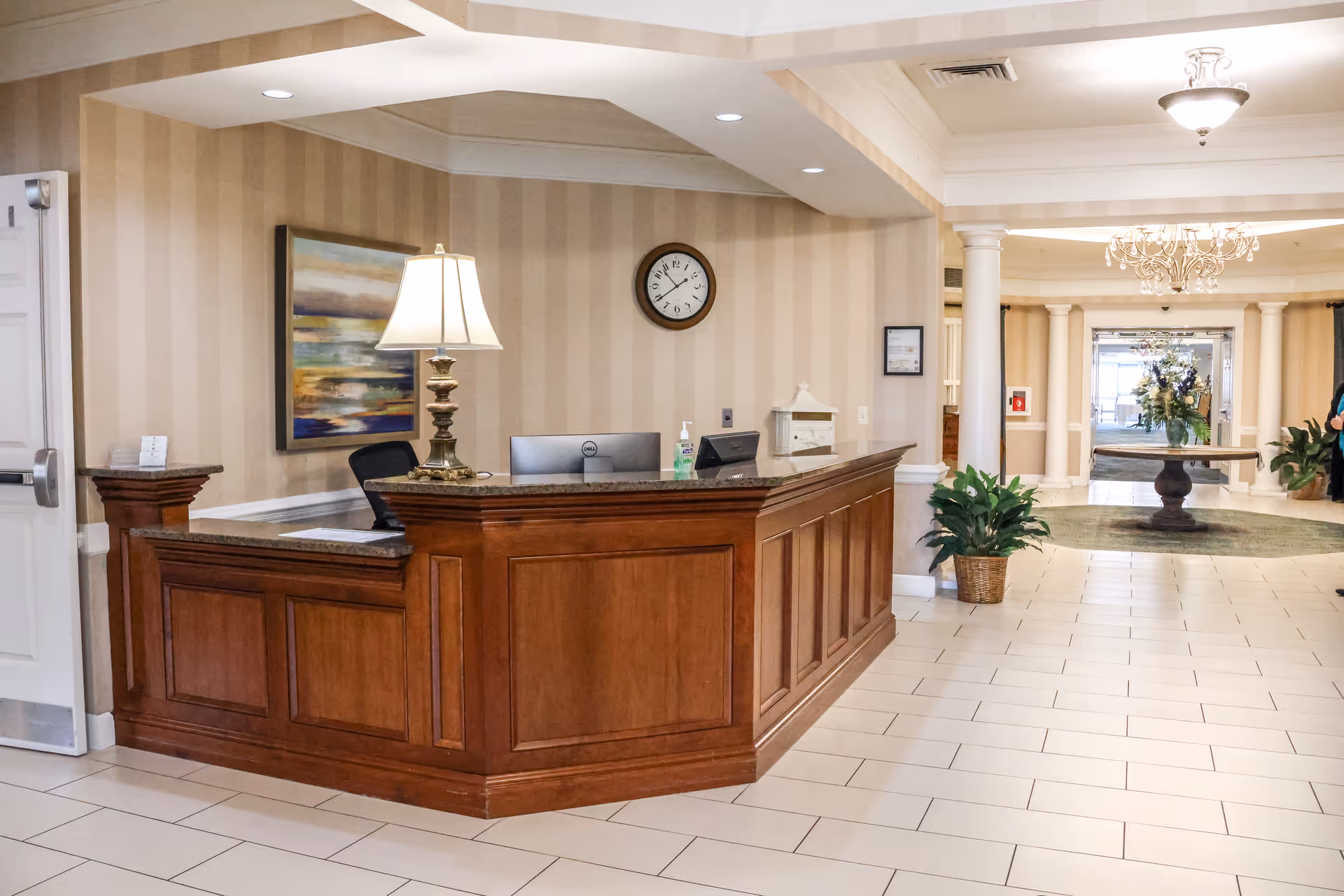 Reception area in a senior living facility with a wooden front desk, a table lamp, a computer monitor, a wall clock, and a painting on the wall. The background shows a hallway with columns, a chandelier, a round table with a flower arrangement, and potted plants.
