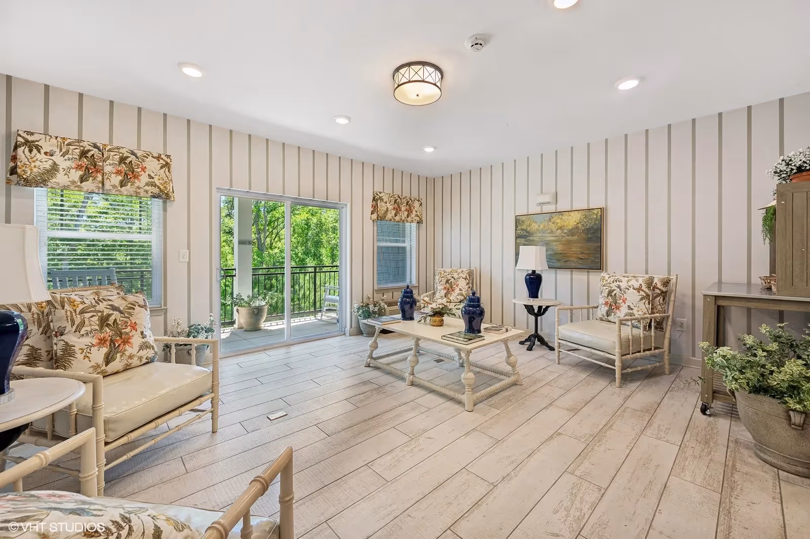 Bright furnished living room with floral-upholstered chairs, a coffee table, and sliding glass doors opening to a balcony.