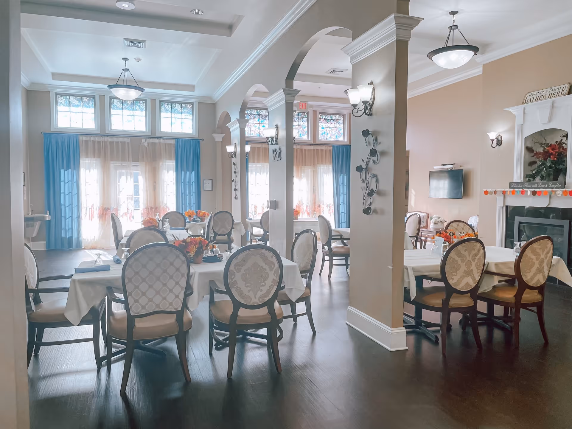 A bright and elegant dining room with several tables covered in white tablecloths, each surrounded by upholstered chairs. The room features large windows with blue and sheer curtains, decorative wall sconces, a fireplace with floral decorations, and a wall-mounted TV. The space has a warm and inviting atmosphere with wooden flooring and arched columns.
