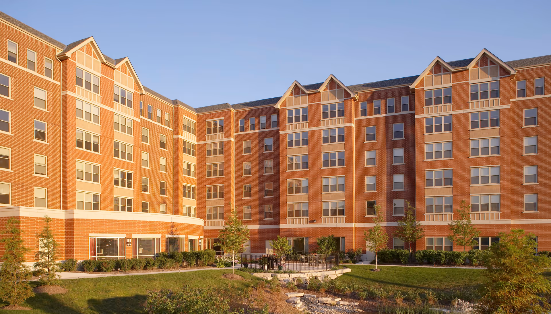 Exterior view of a large multi-story brick building with many windows, surrounded by landscaped greenery and small trees under a clear sky.
