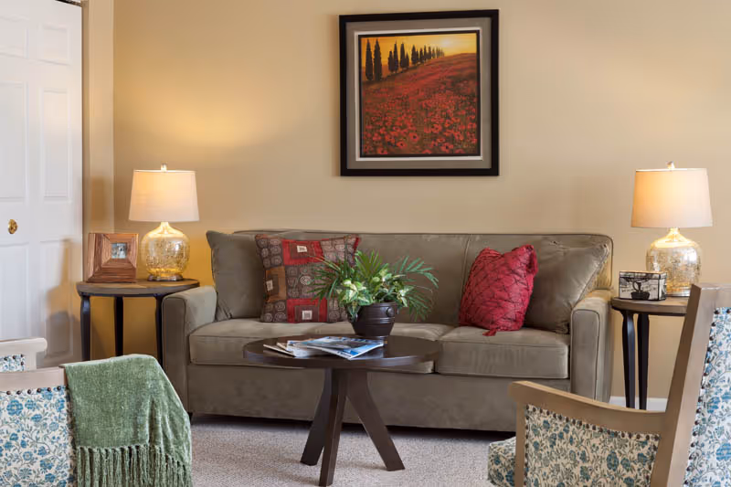 A cozy living room with a beige sofa adorned with patterned and solid red throw pillows. Two side tables with glass lamps flank the sofa. A round wooden coffee table with a potted plant and magazines sits in front of the sofa. Two upholstered chairs with floral patterns and a green throw blanket are partially visible. A framed landscape painting hangs on the beige wall above the sofa.