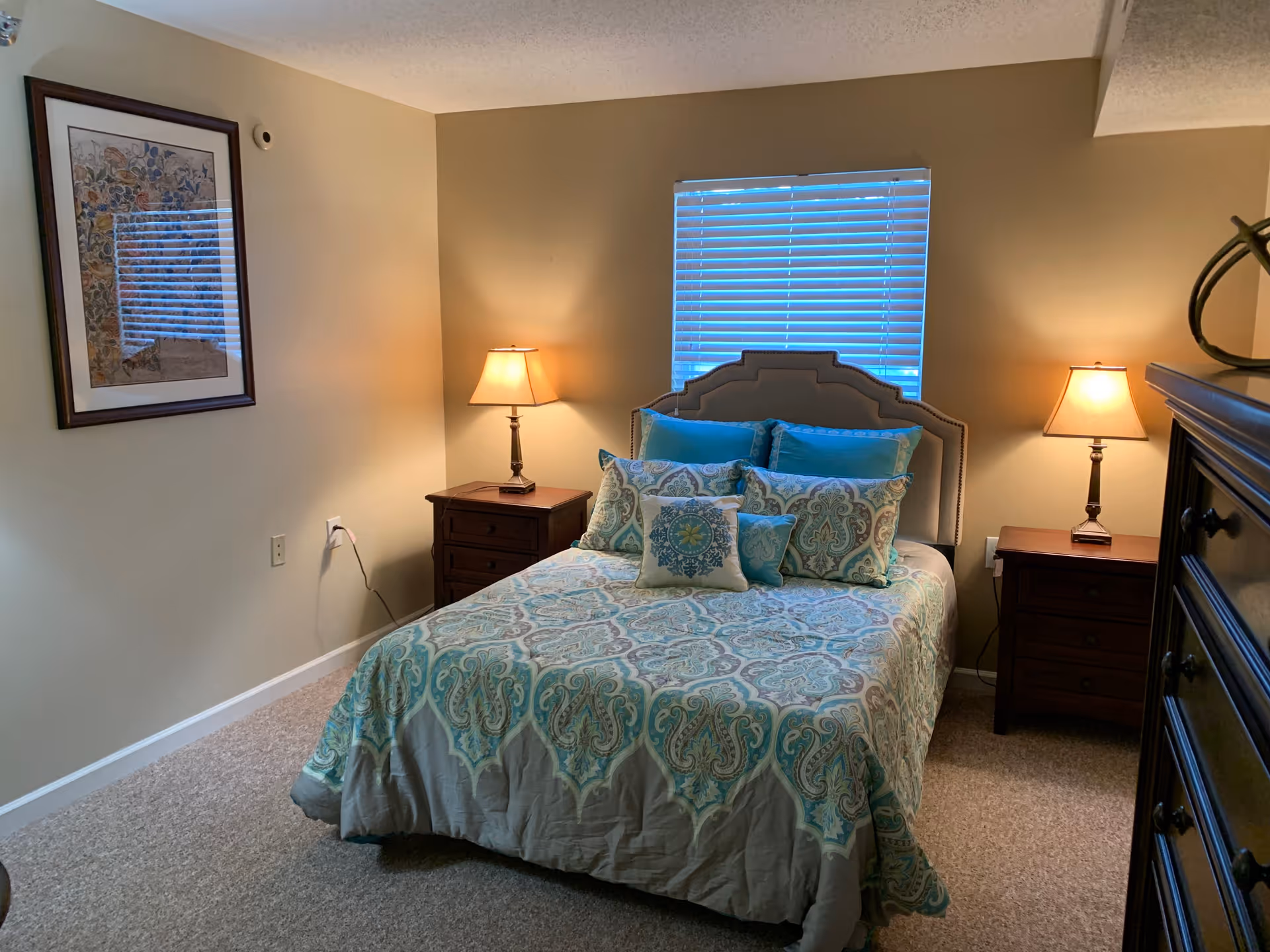 A cozy bedroom with a bed centered against the wall under a window with closed blinds. The bed has a decorative headboard and is made with a patterned comforter and multiple pillows in shades of blue and green. There are two wooden nightstands on either side of the bed, each with a lit table lamp. A framed artwork hangs on the left wall, and a dark wooden dresser is partially visible on the right side of the image.