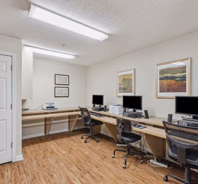 Interior office workspace with desks, computers, chairs, printers, and framed artwork on the walls.