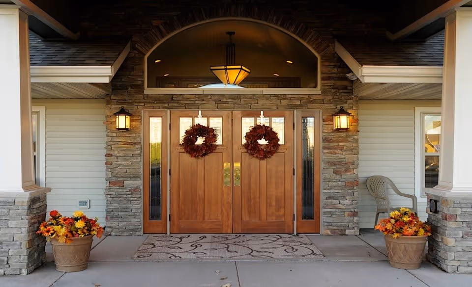 Front entrance of a building with double wooden doors decorated with autumn wreaths. The entrance is framed by stone pillars and siding, with two wall-mounted lantern lights on either side of the doors. There are two large flower pots with fall-colored flowers on each side of the entrance, and a single plastic chair is placed near the right side. A patterned doormat lies in front of the doors.