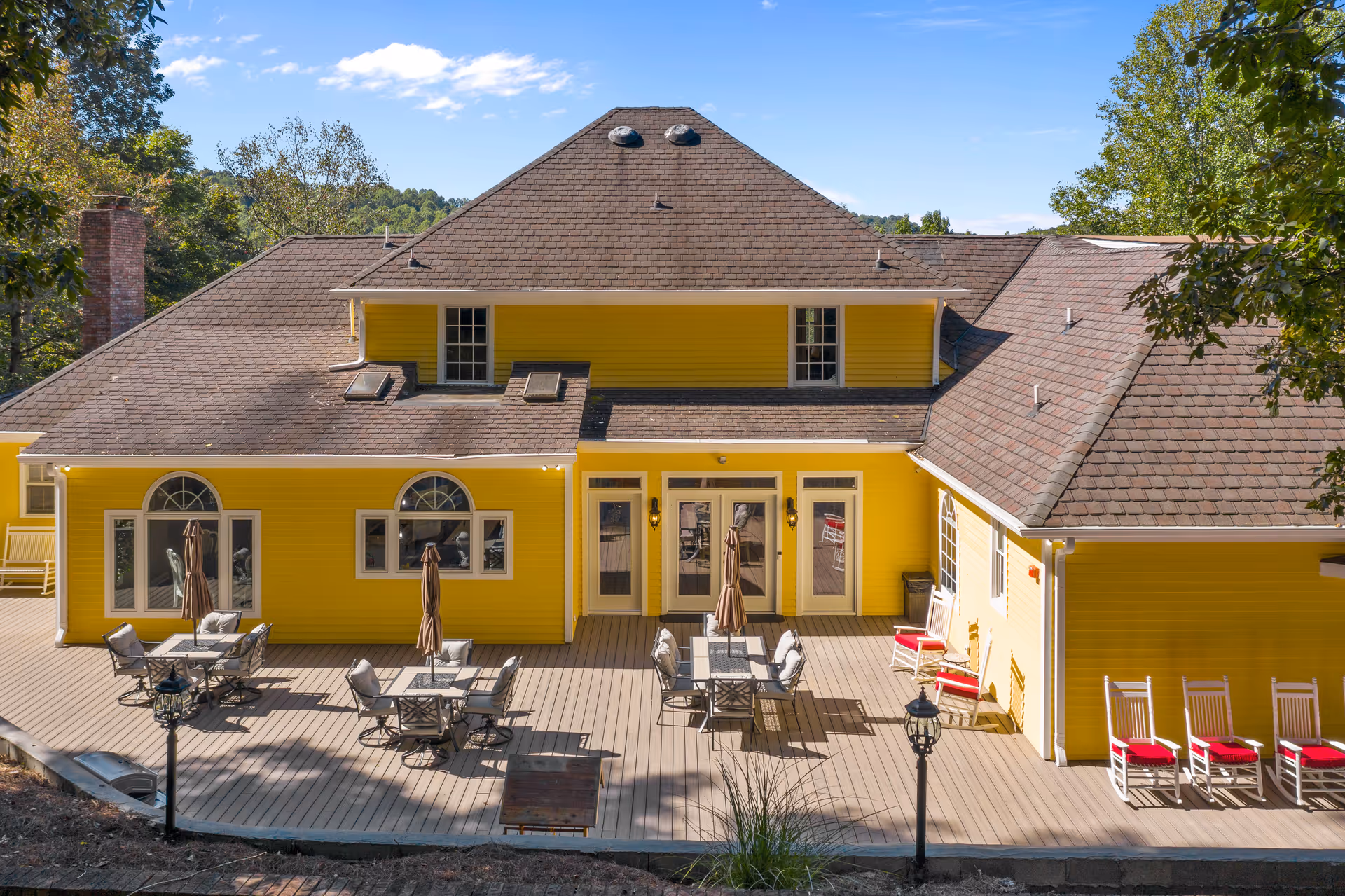 Exterior view of a bright yellow senior living facility building with a large wooden deck featuring multiple tables with umbrellas and cushioned chairs, as well as several white rocking chairs with red cushions. The building has a brown shingled roof and is surrounded by trees under a clear blue sky.