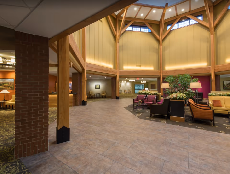 Spacious interior common area with high vaulted ceiling supported by wooden beams, tiled floor, and a seating area with chairs and sofas arranged around small tables. The space is well-lit with lamps and natural light from upper windows, and decorated with plants and artwork on the walls.