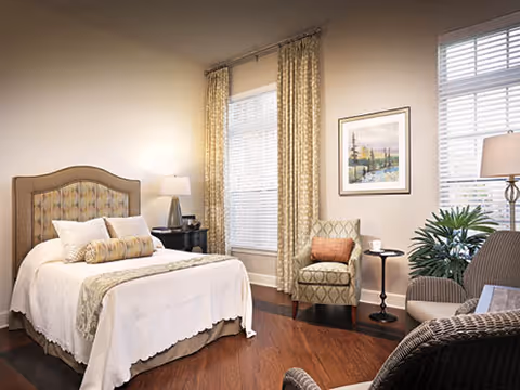 Sunlit bedroom with a bed featuring an upholstered headboard, two armchairs, side tables with lamps, framed art, and large windows with curtains.