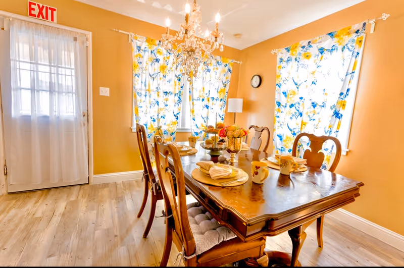 A bright dining room with a wooden dining table set for four, featuring floral-patterned curtains on the windows, a chandelier hanging from the ceiling, and an exit door with a sheer curtain.