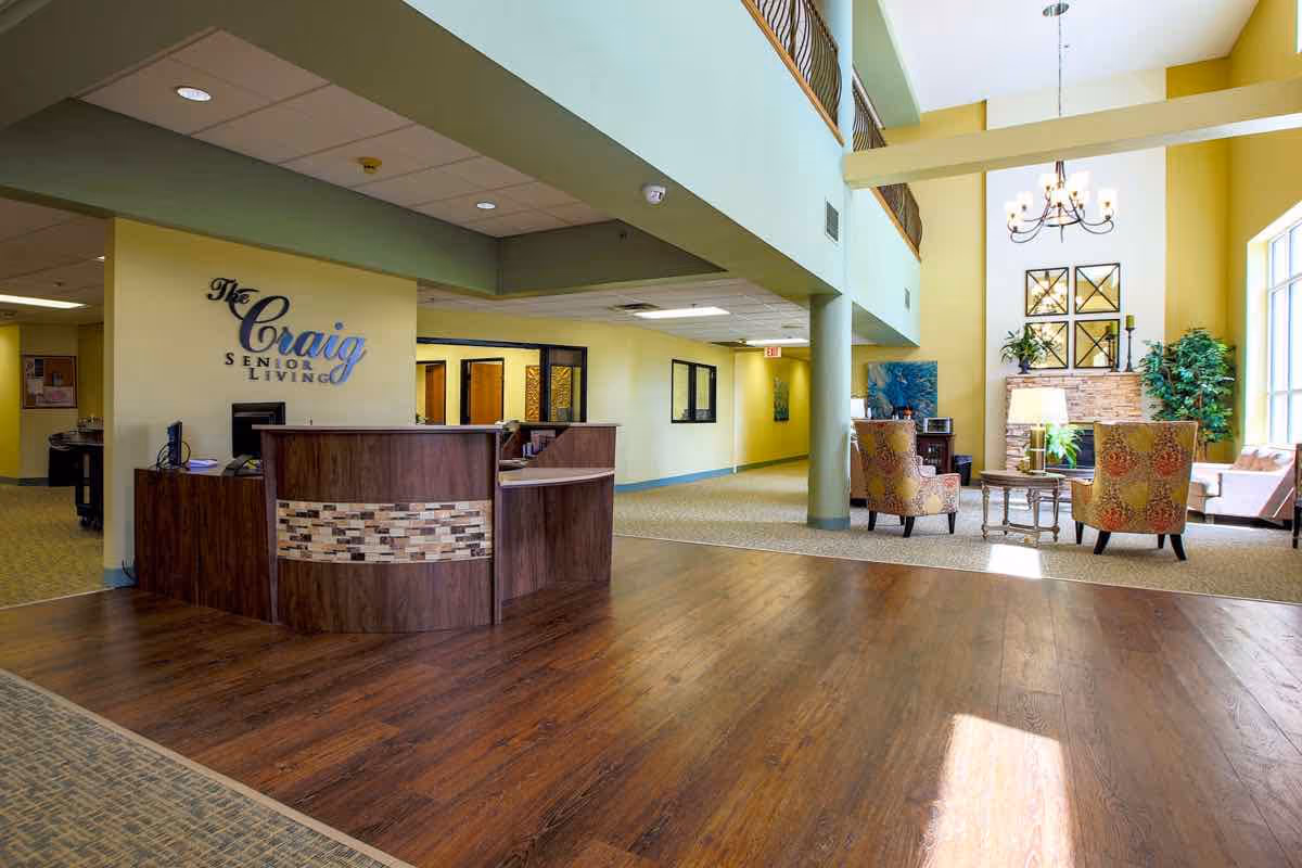 Interior view of The Craig Senior Living facility showing a reception desk with a sign on the wall, a seating area with chairs and a coffee table near a fireplace, large windows letting in natural light, and a high ceiling with a chandelier.