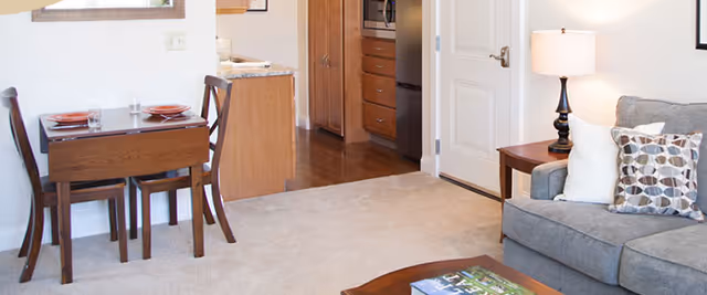A cozy living area featuring a gray sofa with patterned and white pillows, a wooden side table with a lamp, a small dining table set with two chairs and place settings, and a view into a kitchen area with wooden cabinets and a white door.