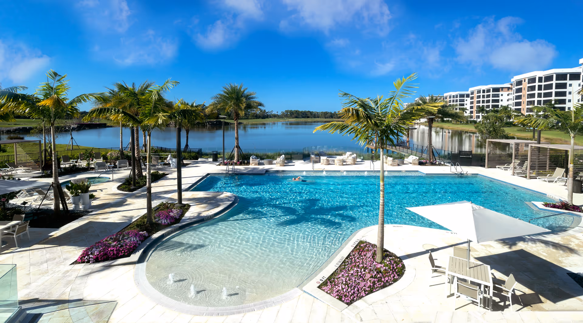 Outdoor swimming pool area with palm trees, lounge chairs, and tables with umbrellas. The pool overlooks a large lake with clear blue skies above. Residential buildings are visible in the background.
