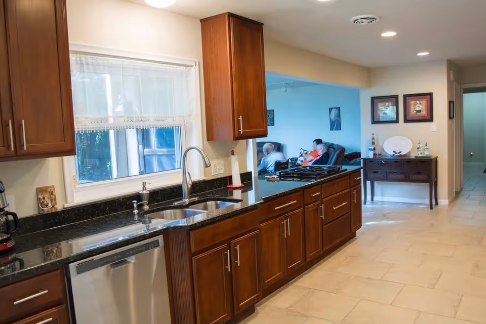 A modern kitchen with dark wooden cabinets, a black granite countertop, a stainless steel dishwasher, and a double sink under a window with white curtains. In the background, a living room area is visible with two elderly people sitting on recliners. The floor is tiled, and the walls are painted in neutral colors with framed artwork and a small table with decorative items.