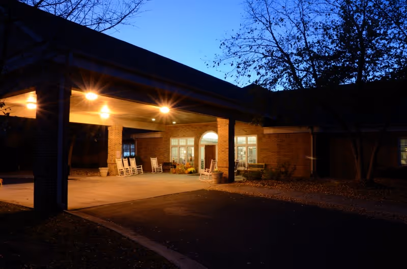 Covered entrance of a senior living facility at dusk with an illuminated canopy, rocking chairs on the porch, and a brick facade.