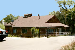 Single-story wood-clad building with a pitched shingle roof, fenced front porch, and surrounding trees viewed from the driveway.