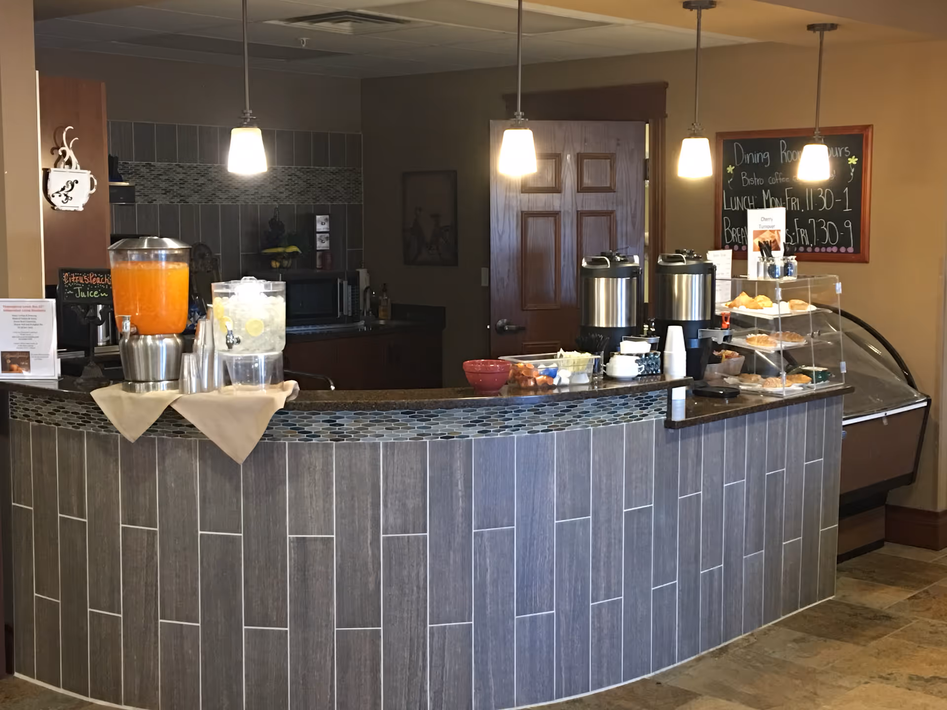 Curved serving counter with beverage dispensers, pastries, cups and pendant lights in a dining area.