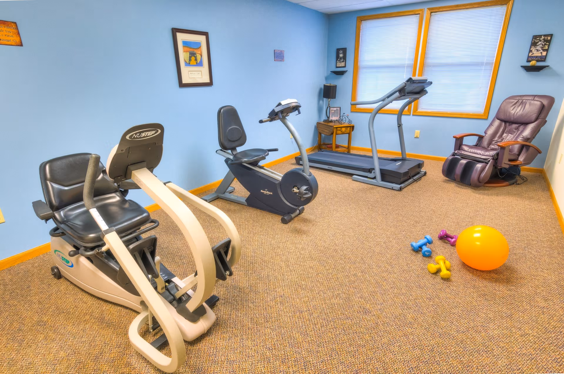 A small exercise room with light blue walls and beige carpet, containing a recumbent exercise bike, a treadmill, a massage chair, and small colorful dumbbells and an orange exercise ball on the floor. There are two windows with blinds and framed pictures on the walls.