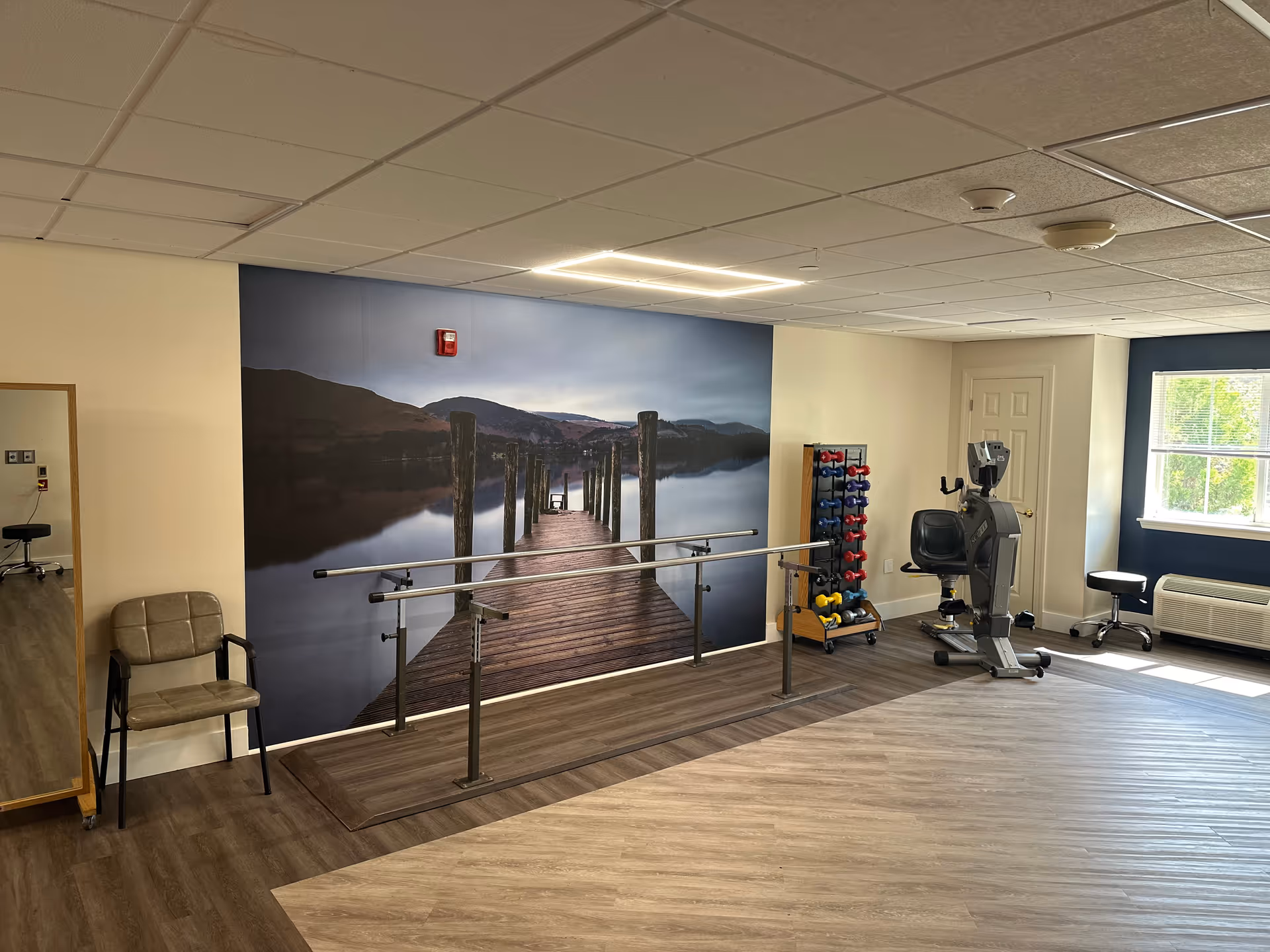 A rehabilitation or exercise room with a large wall mural of a wooden dock extending into a calm lake surrounded by hills. The room has parallel bars for walking exercises, a rack of colorful dumbbells, an exercise bike, a chair, a large mirror, and a window letting in natural light.