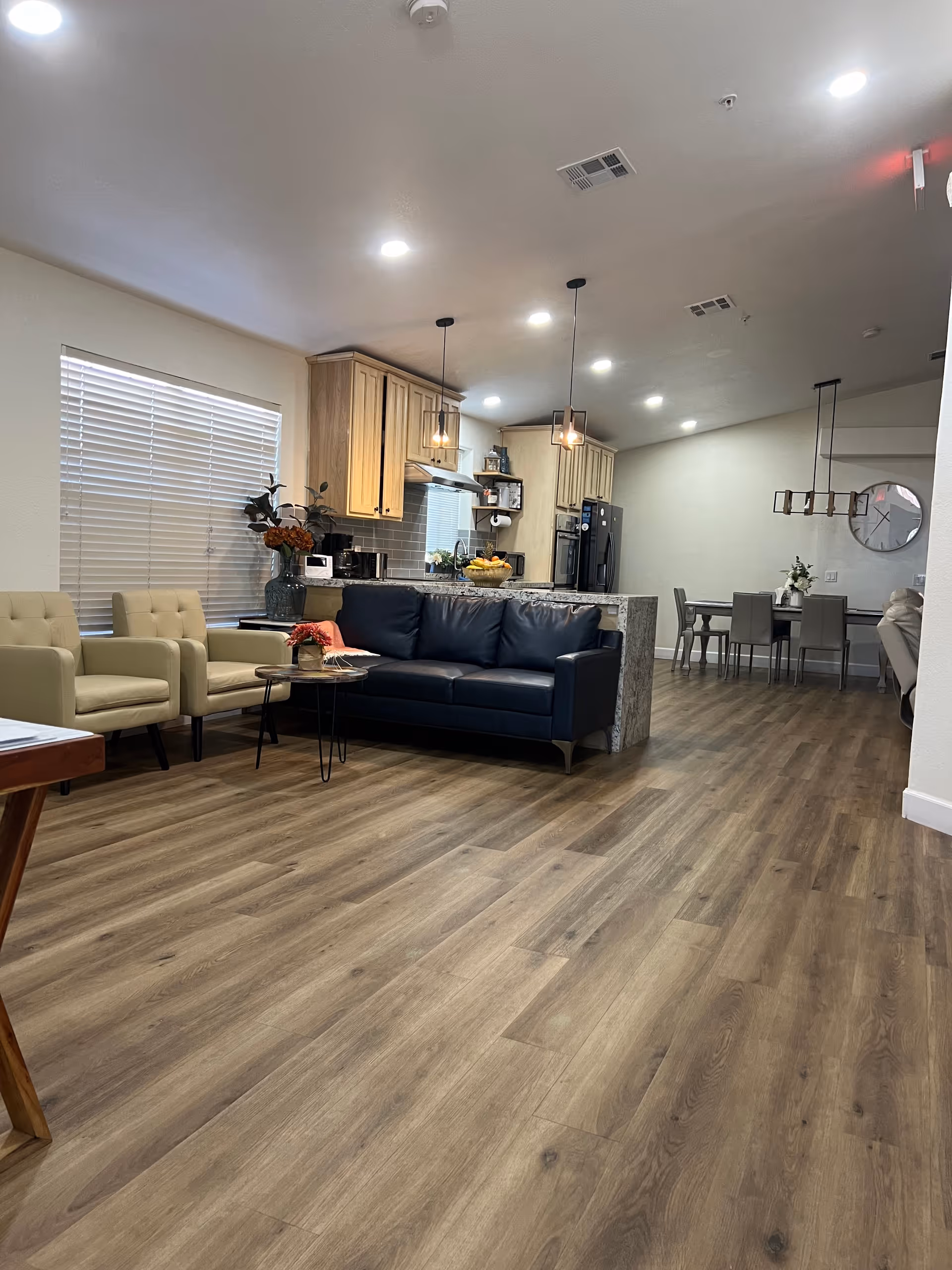 A spacious and modern interior living area with wood flooring, featuring a black leather sofa, two beige armchairs, a small round coffee table with a flower vase, and a kitchen area with wooden cabinets, pendant lights, and a refrigerator. In the background, there is a dining table with chairs and a large wall clock.