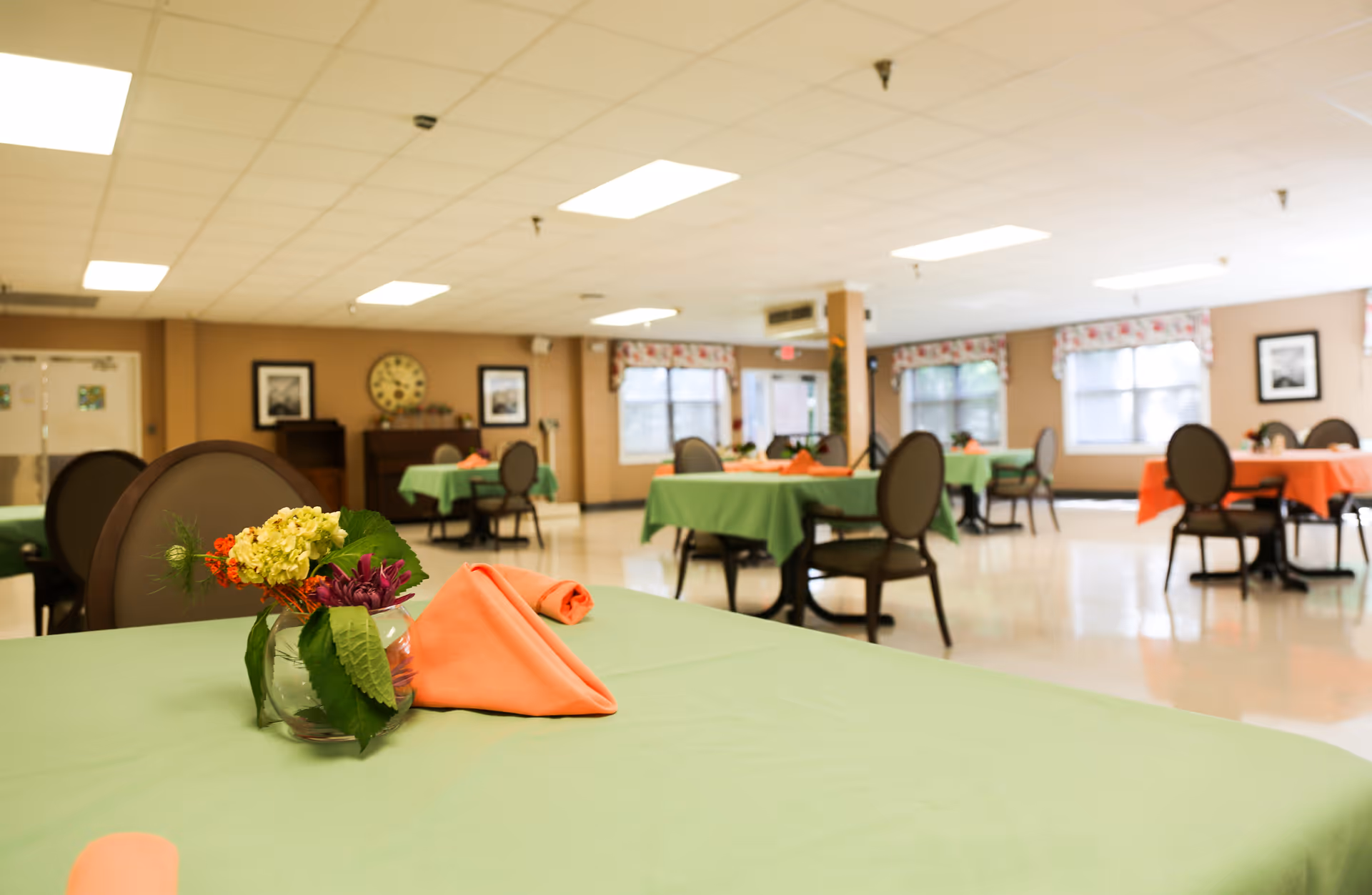 A spacious dining room with multiple tables covered in green and orange tablecloths, each set with folded orange napkins and small floral centerpieces. The room has large windows with floral valances, beige walls, and a clock on the far wall above a piano.