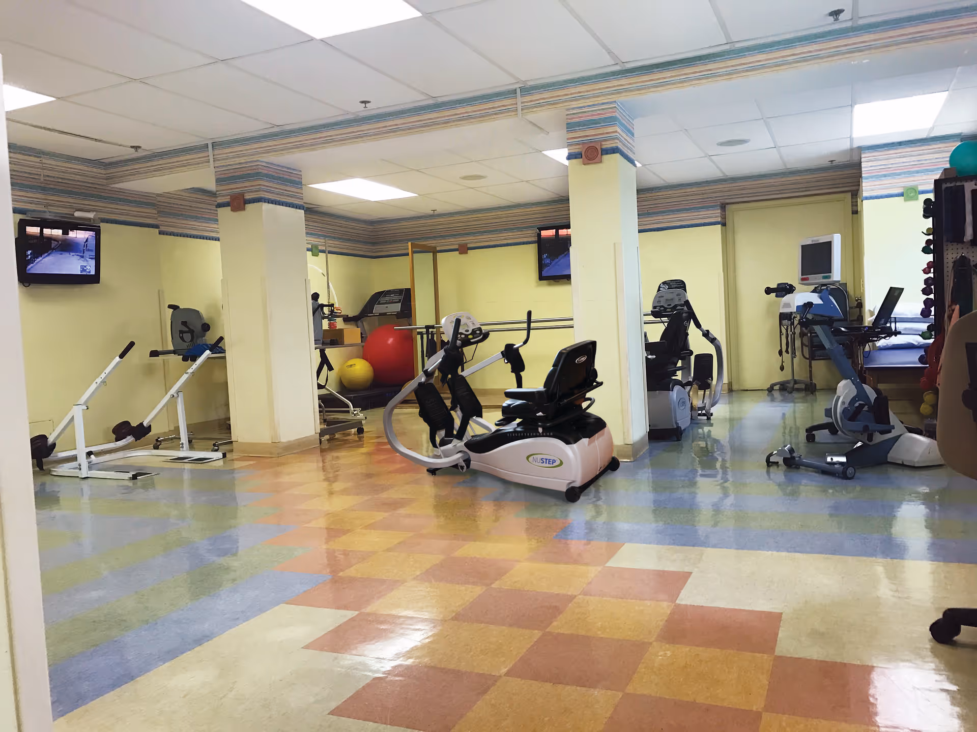 A spacious room with exercise equipment including stationary bikes, a treadmill, and various fitness balls. The room has a colorful checkered floor, light yellow walls with striped wallpaper borders near the ceiling, and two flat-screen TVs mounted on the walls. There are also medical monitoring devices and weights visible in the background.