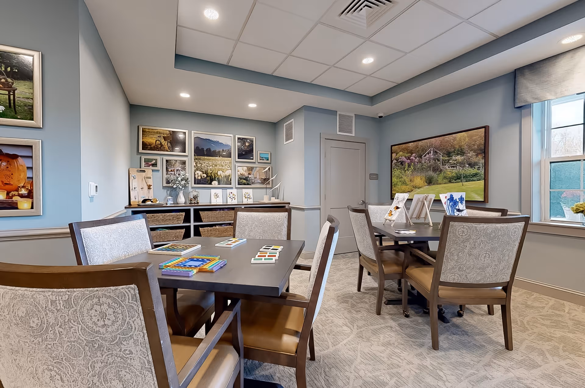 A well-lit activity room with two tables surrounded by cushioned chairs. On the tables are various art supplies and small paintings on display. The walls are decorated with framed landscape photographs and artwork. A window with a valance lets in natural light, and a cabinet with baskets and decorative items is against one wall.