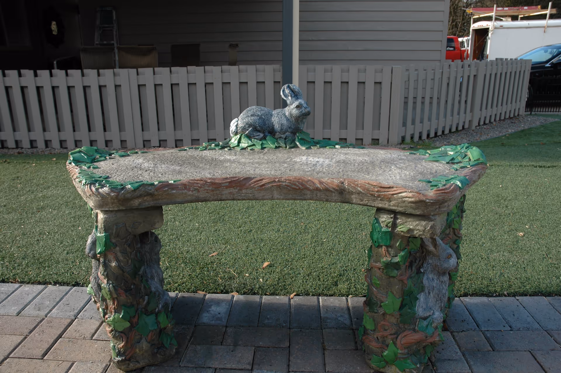 Decorative stone bench topped with a rabbit statue on a paved patio and artificial grass lawn in front of a fenced building.