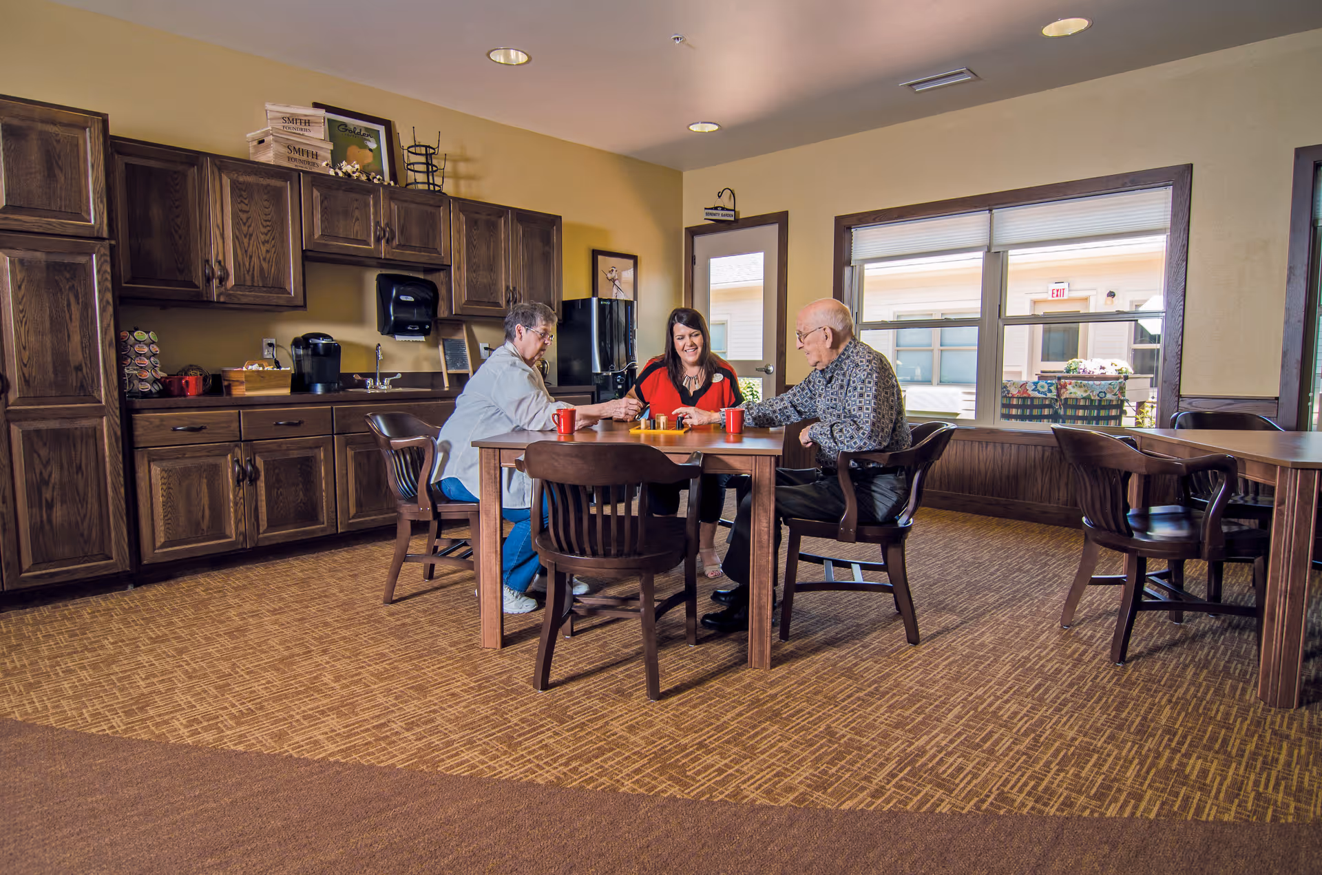 Three people sitting around a wooden table in a room with wooden cabinets and a kitchenette area. They appear to be engaged in a friendly activity or conversation. The room has large windows letting in natural light and carpeted flooring.
