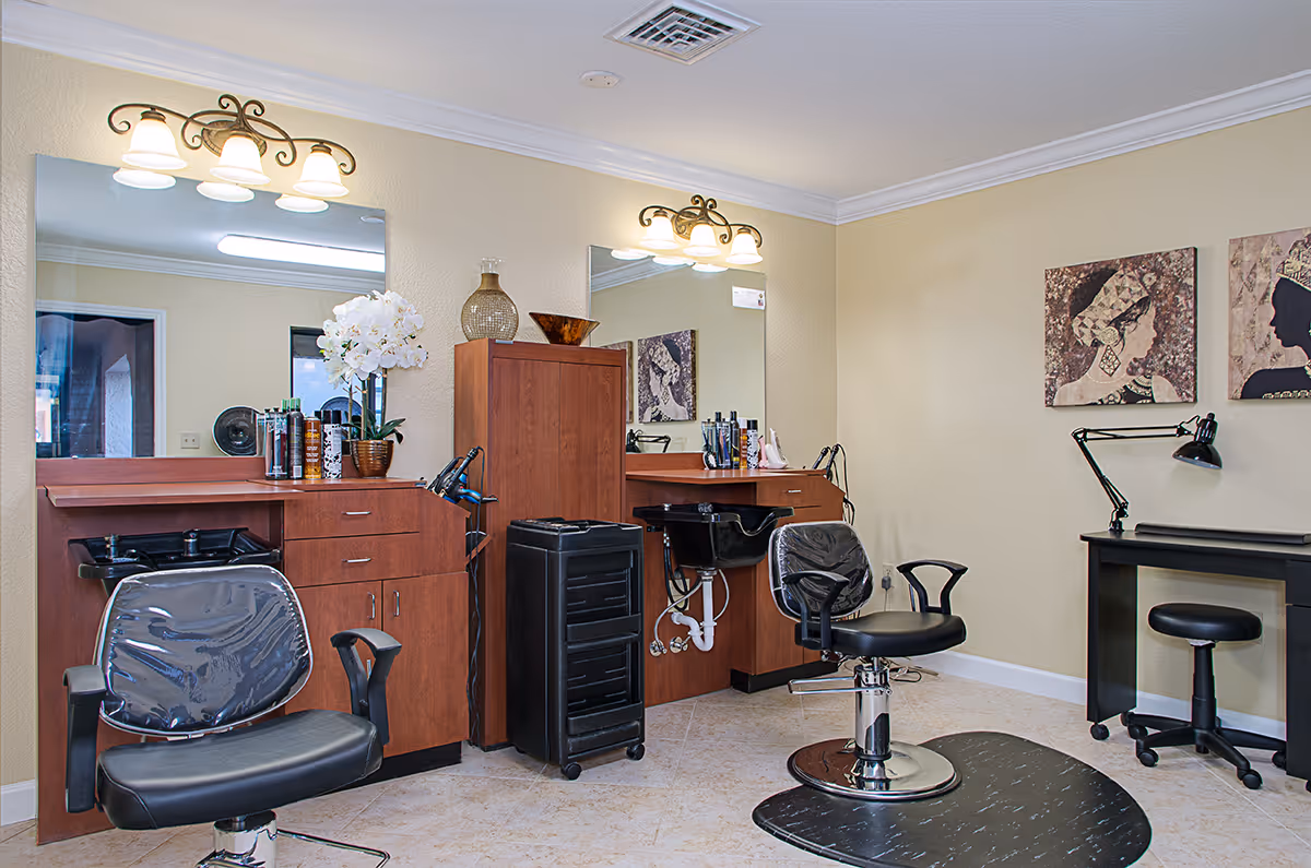 Interior view of a salon area in a senior living facility with two styling chairs in front of large mirrors, wooden cabinets, hair care products, and a small desk with a lamp and stool. The walls are light yellow with decorative artwork.