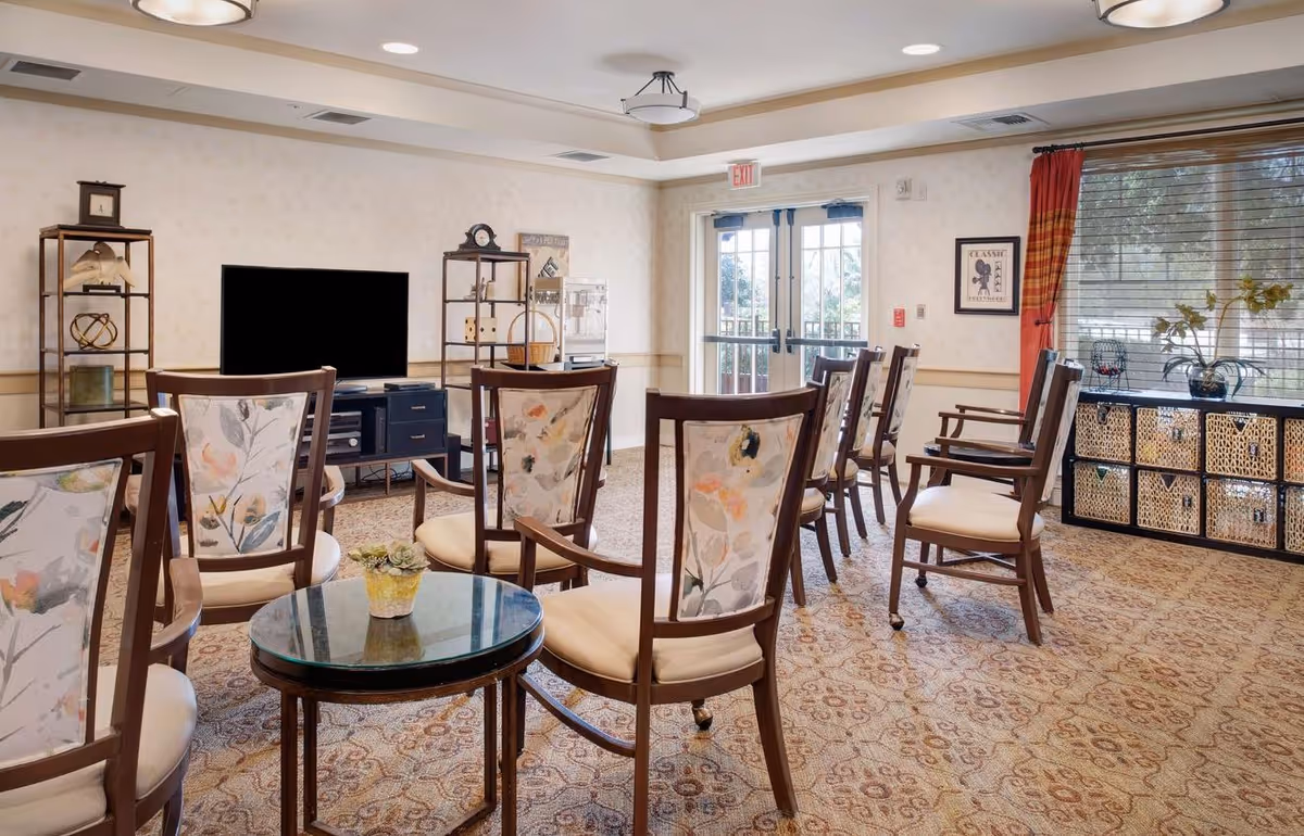 A well-lit communal seating area with rows of floral-upholstered chairs facing a television and glass double doors.