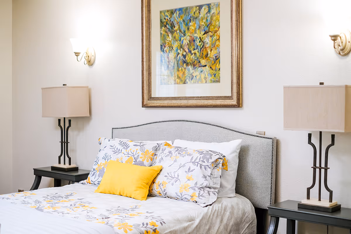 A neatly made bed with floral patterned bedding in shades of yellow and gray, flanked by two black nightstands each with a beige lamp. Above the bed hangs a framed abstract painting with yellow and blue tones. The walls are light-colored and there are two wall sconces providing soft lighting.