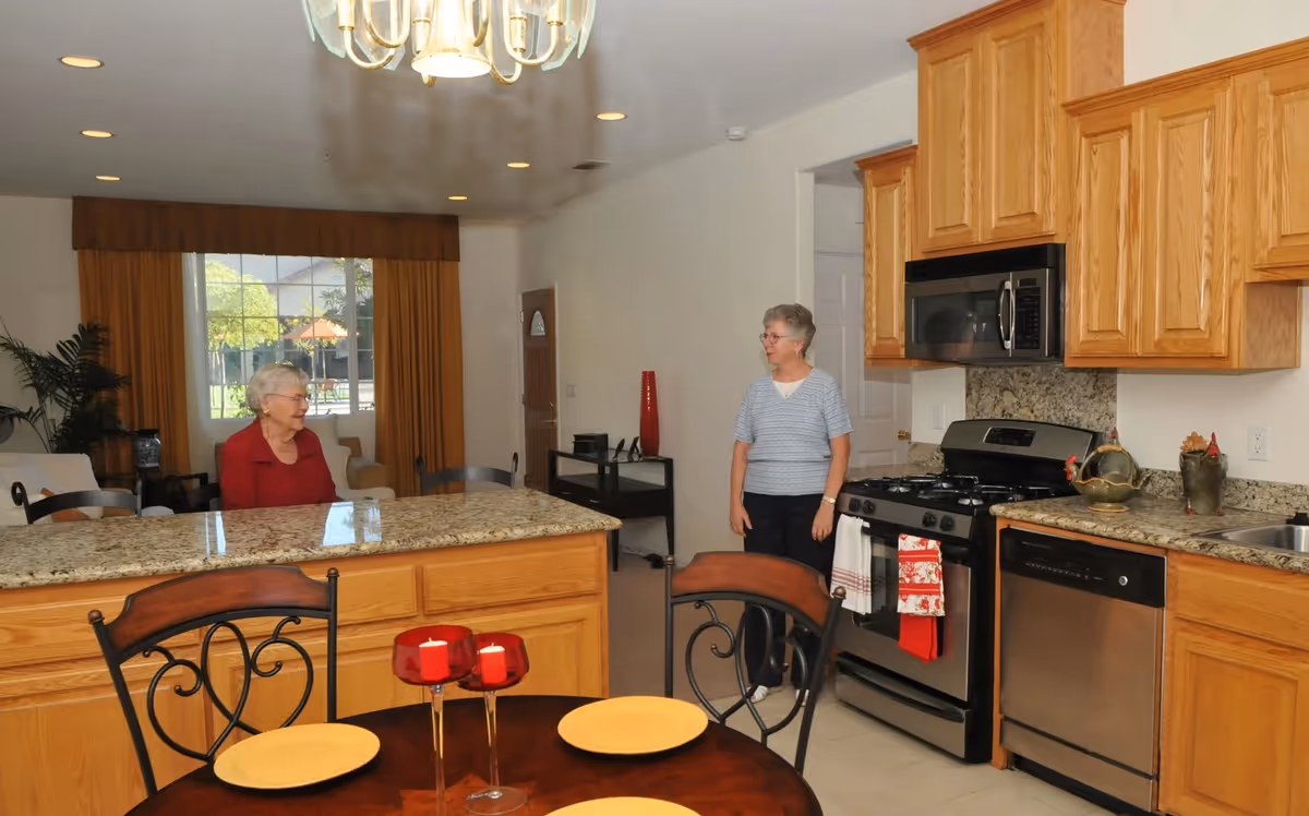 Interior view of a kitchen and living area in a retirement center. Two elderly women are present; one is sitting behind a granite kitchen island, and the other is standing near the stove. The kitchen has wooden cabinets, stainless steel appliances including a stove, microwave, and dishwasher. A round dining table with yellow plates and red candles is in the foreground. The living area in the background has a window with yellow curtains and some furniture.