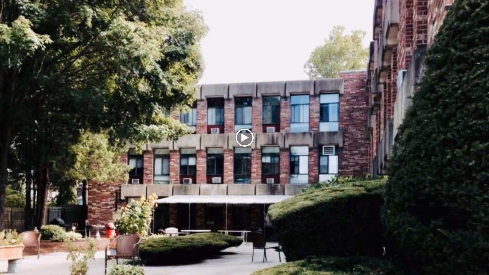 Brick three-story rehabilitation building with a shaded outdoor courtyard, patio chairs, and trimmed bushes.