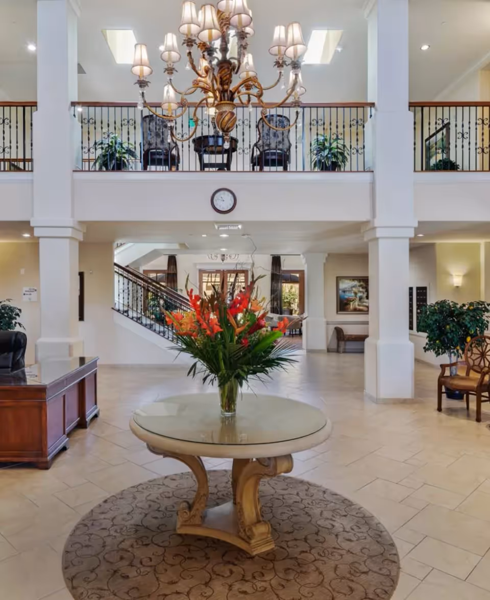 Spacious and elegant senior living facility lobby with a round table in the center holding a large vase of vibrant red and orange flowers. The area features tiled flooring, a patterned round rug, white pillars, a wooden desk on the left, and a staircase with wrought iron railings leading to an upper balcony with chairs and plants. A large chandelier hangs from the ceiling, and a clock is mounted on the wall above the entrance to the staircase.