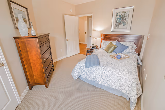 Furnished bedroom with a patterned bed, wooden headboard and dresser, bedside table and an open door to a hallway.